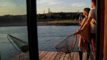 Paula y Ezequiel Naumiec en el deck de una de las suites de la bodega Trina, en Río Colorado, que da al río Colorado. Con copa en mano, el atardecer en esta naturaleza es una experiencia enriquecedora. 